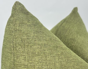 May include: Two olive green throw pillows featuring a textured, woven fabric construction. The pillows have a distinct, visible weave pattern with a slightly raised surface, showcasing a consistent, solid green color throughout the material.