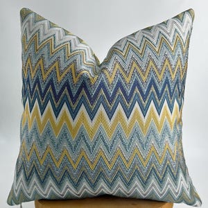 May include: Square decorative pillow with a zig-zag pattern in shades of blue, yellow, and gray. The pillow has a textured appearance and is likely made of woven fabric. The design is a repeating chevron pattern, adding a modern touch to home decor.