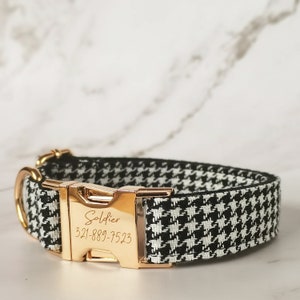 May include: Black and white houndstooth dog collar with a gold buckle. The buckle is engraved with the text "Soldier" and a phone number.