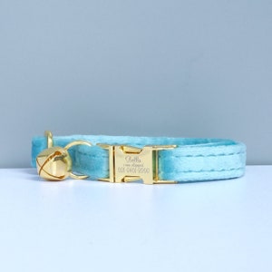 Puede incluir: Un collar de gato de terciopelo azul claro con una hebilla dorada y una campanilla. La hebilla est&aacute; grabada con el nombre "Bella" y el texto "I am chipped 021-6401-8996".