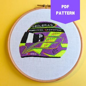 Op de afbeelding: Borduurwerk van een paarse en groene racehelm met de tekst "SCLERAF CROWDSTRIKE CROWDSTRIKE" aan de zijkant. De helm bevindt zich in een houten borduurring.