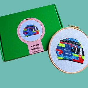 May include: A green box with a pink circle containing a colourful embroidered helmet. The box says "Oscar Piastri Embroidery Kit". The helmet is also embroidered on a white fabric in a wooden embroidery hoop.