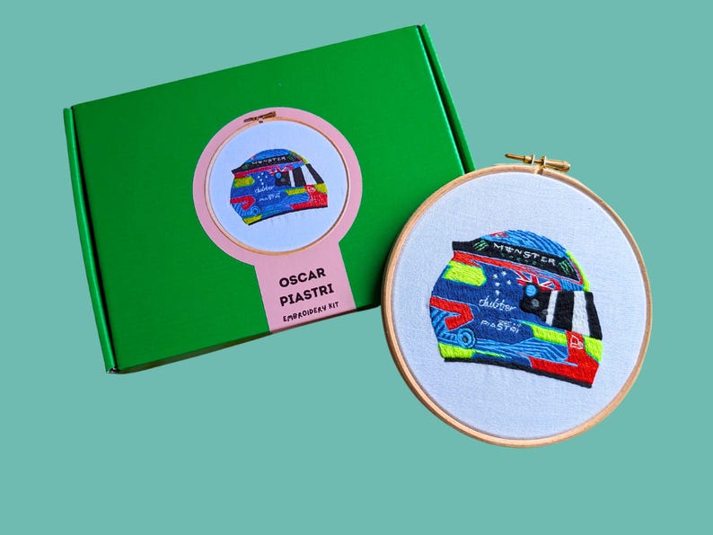 May include: A green box with a pink circle containing a colourful embroidered helmet. The box says "Oscar Piastri Embroidery Kit". The helmet is also embroidered on a white fabric in a wooden embroidery hoop.