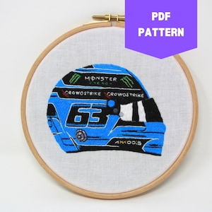 Op de afbeelding: Borduurwerk van een blauwe racehelm met nummer 63, Monster Energy logo en Crowstrike branding. De helm bevindt zich op een witte stoffen achtergrond in een houten borduurring.