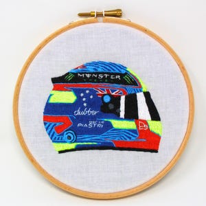 May include: Embroidered racing helmet with a blue, green, red and white design. The helmet has the words "Monster Energy" and "Oscar Piastri" on it.