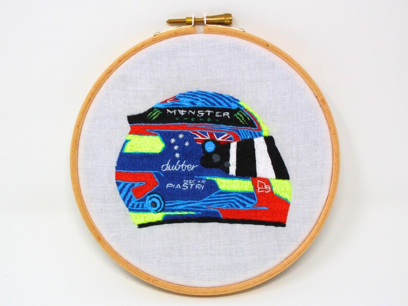 May include: Embroidered racing helmet with a blue, green, red and white design. The helmet has the words "Monster Energy" and "Oscar Piastri" on it.