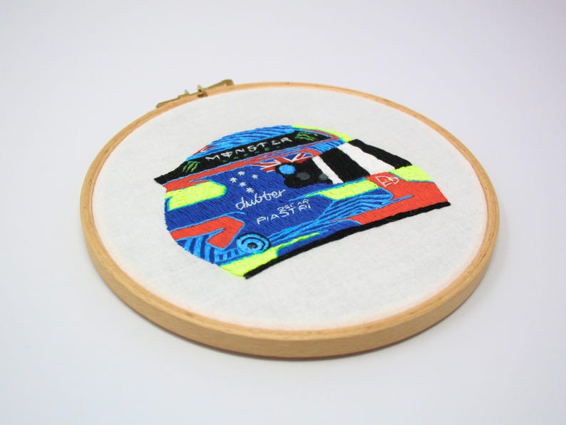 May include: Embroidered design of a racing helmet with a blue, red, yellow and black colour scheme. The helmet has the words "Monster" and "Piastri" on it.