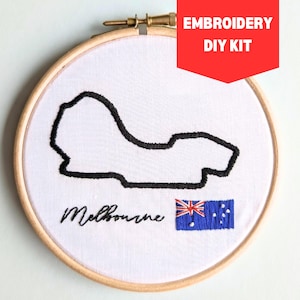 Puede incluir: Kit de bordado con un contorno negro bordado del circuito de Melbourne sobre tela blanca. La palabra "Melbourne" está bordada en escritura negra debajo del contorno de la pista, con una bandera australiana. El bordado está en un aro de madera.