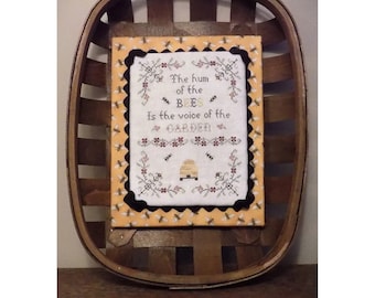 May include: A framed cross-stitch embroidery with a yellow background and black border. The embroidery features a beehive, bees, and the text "The hum of the bees is the voice of the garden."