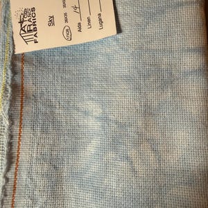 May include: Sky-colored fabric with a woven texture, featuring a label with the text "Atomic Ranch Fabrics." The label also indicates dimensions of 26x35 inches and 17x26 inches, along with the word "Aida" and the number "14."