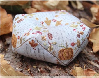 May include: A cross-stitch biscornu pillow with a fall theme, featuring pumpkins, leaves, and berries. The text "Heartstring Samplery" and "Gift of October Biscornu" are visible.