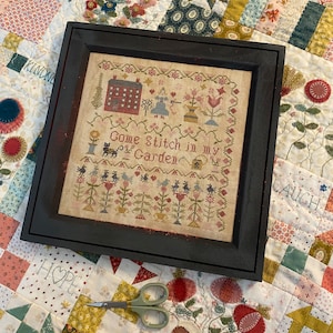 Può includere: Un ricamo a punto croce incorniciato con una cornice nera e uno sfondo crema. Il ricamo presenta una casa, fiori, uccelli e il testo "Come Stitch in my Garden".