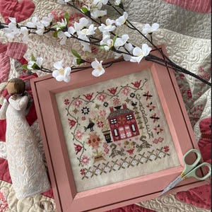 Könnte beinhalten: Eine gerahmte Kreuzstich-Stickerei mit einem roten Haus mit einem weißen Schornstein und einer Gartenszene. Der Rahmen ist hellrosa und die Stickerei ist auf einem cremefarbenen Stoff. Der Text "GARDEN" ist in der Stickerei sichtbar.