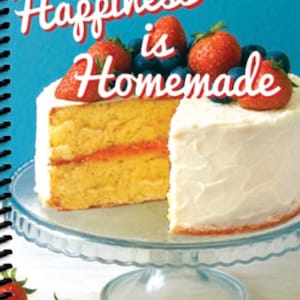 May include: A cookbook titled "2026 Nashville Needlework Market Cookbook" with the text "Happiness is Homemade." A slice of cake with white icing and strawberries sits on a glass cake stand. The cookbook has a blue background.