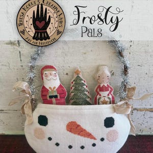 Può includere: Un cestino fatto a mano a forma di pupazzo di neve pieno di figure natalizie cucite. Il cestino ha un naso di carota, occhi di carbone nero e una bocca sorridente. Il cestino contiene un Babbo Natale, un albero di Natale e un elfo. Il testo "Frosty Pals" è sopra il cestino.
