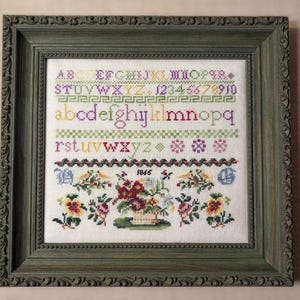 May include: Framed cross-stitch sampler with a green ornate frame. The sampler features the alphabet, numbers, and floral designs in various colors. The year "1865" is stitched below a floral basket.