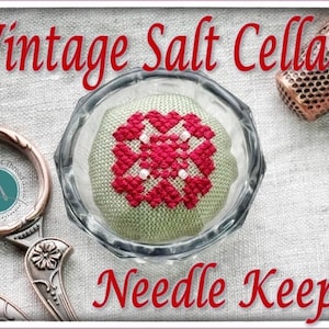 Könnte beinhalten: Ein Vintage-Nadelkissen mit dem Text "Vintage Salt Cellar Needle Keep". Der transparente Glasbehälter enthält ein grünes Kissen mit einem roten Kreuzstichmuster. Ein kupferner Fingerhut und eine Schere sind ebenfalls sichtbar.