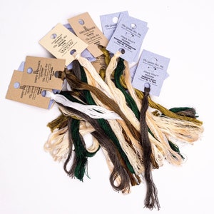 May include: A collection of embroidery floss skeins in shades of green, brown, and beige. The skeins are labeled with the brand name "The Gentle Art" and the color names "Garden Gate", "Roasted Marshmallow", and "Oatmeal".