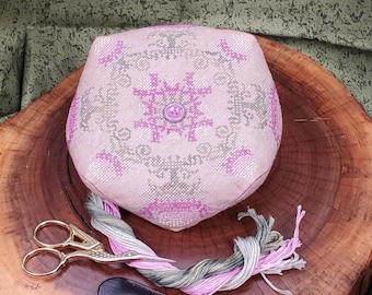 May include: A pink and gray needlepoint pincushion with a floral design and a single button in the center. The pincushion is sitting on a wooden surface with a pair of gold scissors, a leather needle case, and a skein of pink and green embroidery floss.