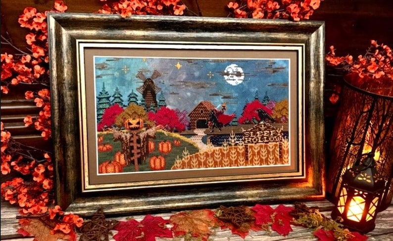 K&ouml;nnte beinhalten: Ein gerahmtes Kreuzstichbild, das eine Szene aus Sleepy Hollow mit einer Vogelscheuche mit K&uuml;rbiskopf, einer Windm&uuml;hle, einer &uuml;berdachten Br&uuml;cke und einem Reiter unter einem Vollmond zeigt. Der Text Sleepy Hollow ist unten rechts eingestickt.