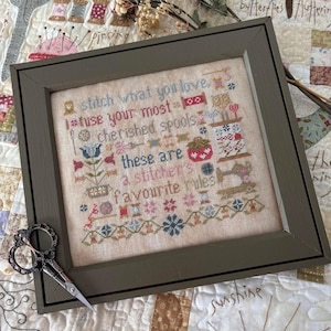 Może przedstawiać: Oprawiony haft krzyżykowy z napisem "stitch what you love, use your most cherished spools, these are a stitcher's favourite rules." Rama jest oliwkowozielona. Para srebrnych nożyczek spoczywa na rogu ramy.