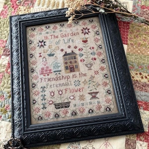 Könnte beinhalten: Gerahmtes Kreuzstich-Kunstwerk mit dem Text "In The Garden of Life. Friendship is the Perennial Flower." Der schwarze Rahmen umgibt ein detailliertes, farbenfrohes Design mit einem Haus, Blumen und Figuren. Eine Schere liegt unten.