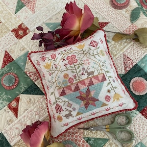 Può includere: Un cuscino bianco con un ricamo a punto croce floreale rosso, rosa e verde. Il cuscino è su una trapunta bianca e verde con un motivo a stella. Ci sono rose secche sulla trapunta.