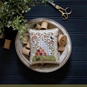 May include: A white cross-stitch sampler with a house, a bird, and tomatoes on a vine. The sampler is in a wooden bowl with spools of thread. The text "Tomato Hill" and "Plum Street Samplers" is below the image.