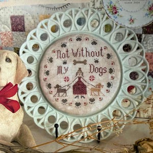 Può includere: Un piatto decorativo con un bordo traforato azzurro presenta un disegno a punto croce. Il punto croce recita "Not Without My Dogs" con sagome di cani. Un piccolo piatto floreale è sullo sfondo. Il piatto è esposto su un supporto nero.