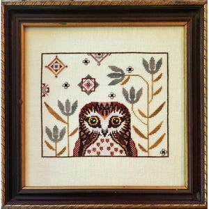 May include: A cross-stitch embroidery of an owl with brown and pink feathers, surrounded by stylized plants and flowers in shades of blue, brown, and gold. The owl has large, round eyes and a small beak.