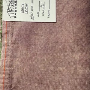 May include: A piece of fabric in a muted mauve color, with a tag that reads "Cherry Cordial" and dimensions of 17x26 inches. The fabric has a textured appearance and a green border. The tag also indicates the fabric is linen.