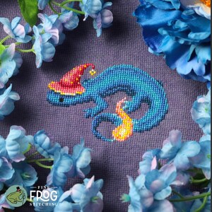 May include: Cross-stitch design of a blue chameleon wearing a red wizard hat with a yellow star. The chameleon has a flame-like tail and is set against a purple background, surrounded by blue flowers. The text "FINE FROG STITCHING" is at the bottom.