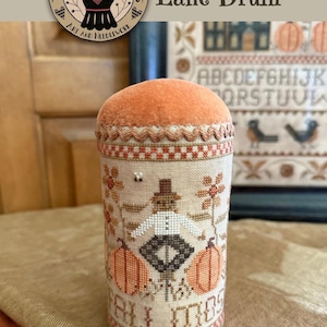 May include: A cylindrical drum-shaped pincushion featuring a cross-stitch design of a scarecrow standing between two orange pumpkins. The fabric has a beige background with brown and orange accents, topped with a solid orange felt cap and decorative trim.