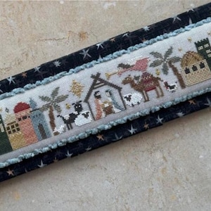 May include: A cross-stitch design showing a nativity scene with buildings, palm trees, and animals. The design has a light blue border and is set against a dark blue fabric with white stars.