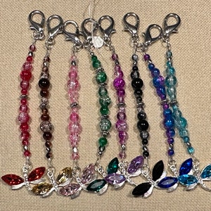 May include: A selection of beaded dragonfly charms in shades of red, brown, pink, green, purple, black, and blue. Each charm has a silver-coloured clasp and a dragonfly pendant with crystal details. The beads are of different sizes and textures.