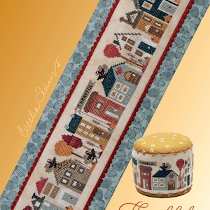 May include: Cross stitch pattern featuring a row of houses with a Thanksgiving theme. The pattern includes a turkey, pumpkins, and the word "Thankful". The pattern is titled "Thankful Tiny Town".