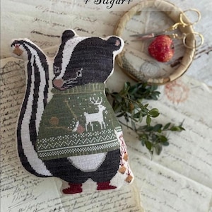 May include: A handcrafted skunk plushie from the "Animal Cracker Series" wearing a green sweater with a deer design. The skunk has black and white stripes and red boots. The image includes the text "Animal Cracker Series + Sugar+" and "Design by Stacy Nash".