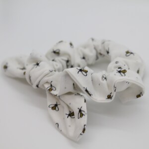 May include: White scrunchie with a bee pattern. The scrunchie is made of a soft, stretchy fabric and is tied in a bow.