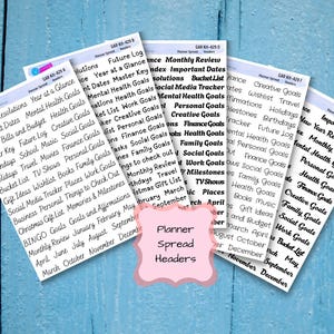 May include: A collection of white planner spread header stickers with black text, including categories like "New Year Resolutions" and "Monthly Review." The stickers are arranged on a blue wooden surface, with a pink label that reads "Planner Spread Headers."