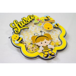 Puede incluir: Una decoración de fiesta de abejas amarilla y negra con el nombre "Luisa" en letras cursivas amarillas. La decoración presenta una niña abeja de dibujos animados con un vestido a rayas amarillo y negro y sosteniendo una flor. La decoración está rodeada de cuentas amarillas y blancas.