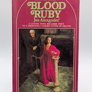 May include: A vintage paperback book titled Blood Ruby by Jan Alexander with a magenta cover. The cover art depicts a person in a long magenta dress standing by a pedestal with a red stone, while a person in a dark suit stands in the background.