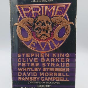 May include: A vintage paperback book titled "Prime Evil" with a black and purple cover. The cover features a skull and horns design in copper color, with the names of horror authors like Stephen King and Clive Barker.