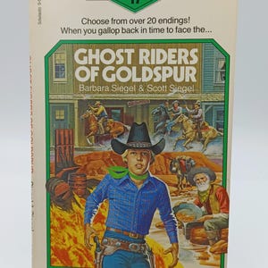 Peut inclure: Un livre de poche intitulé "Ghost Riders of Goldspur" par Barbara Siegel & Scott Siegel. La couverture présente une illustration de cow-boy avec une toile de fond de ville et le texte "Twistaplot 17". Le livre est sur un support blanc.