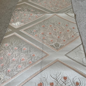 Può includere: Primo piano di un tessuto argento e pesca con un motivo ripetitivo di pavoni. Il tessuto ha una lucentezza discreta e una superficie testurizzata.