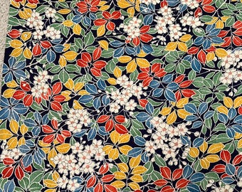 May include: Close-up of a fabric with a repeating floral pattern. The design features white blossoms, red, yellow, green, and blue leaves against a dark navy background. The fabric has a detailed, textured appearance, suggesting a high-quality textile.