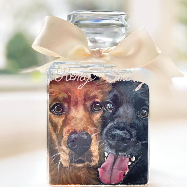 May include: A clear glass bottle with a cream-colored bow tied around the neck. The bottle features a painted portrait of two dogs, one brown and one black. The text "Henry & Silas" is written on the bottle.