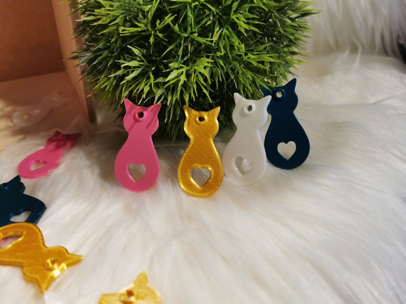 Puede incluir: Cinco encantos con forma de gato de colores vivos con un coraz&oacute;n recortado. Los encantos son rosa, dorado, blanco, azul y negro.