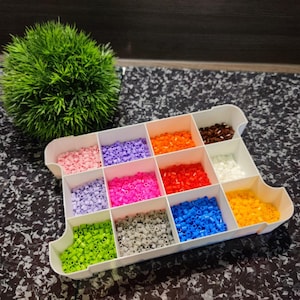 Könnte beinhalten: Ein weißer Plastikbehälter mit 12 Fächern, gefüllt mit bunten Perlen. Die Perlen sind in Reihen und Spalten angeordnet, wobei jedes Fach eine andere Farbe enthält.