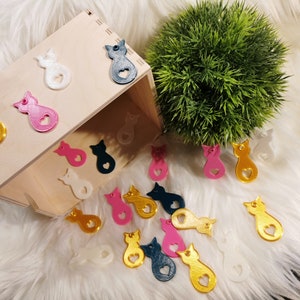 Puede incluir: Una caja de madera llena de dijes con forma de gato de varios colores. Los dijes est&aacute;n hechos de pl&aacute;stico y tienen un coraz&oacute;n cortado en el medio. Los dijes son de varios colores, incluyendo dorado, rosa, blanco y azul.