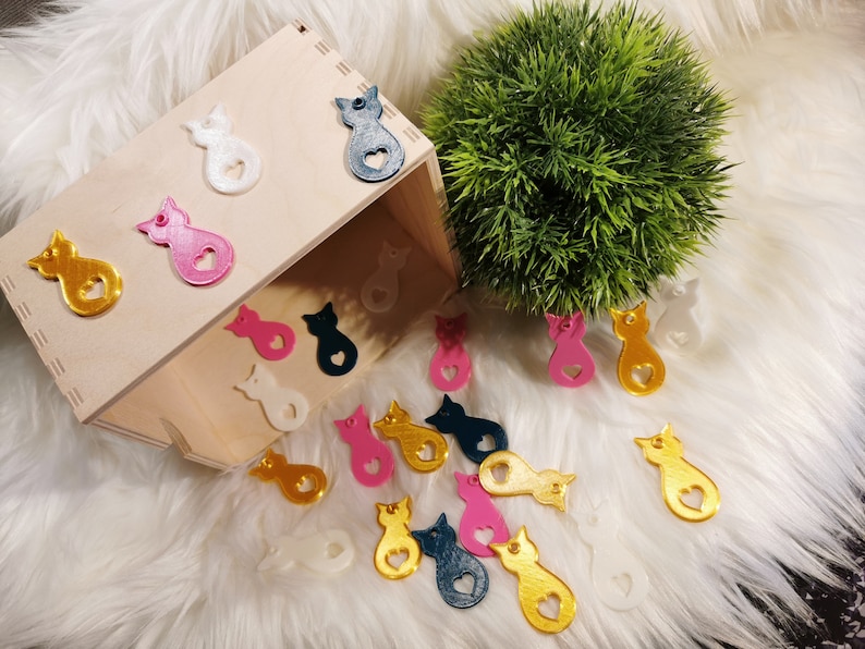 Puede incluir: Una caja de madera llena de dijes con forma de gato de varios colores. Los dijes est&aacute;n hechos de pl&aacute;stico y tienen un coraz&oacute;n cortado en el medio. Los dijes son de varios colores, incluyendo dorado, rosa, blanco y azul.